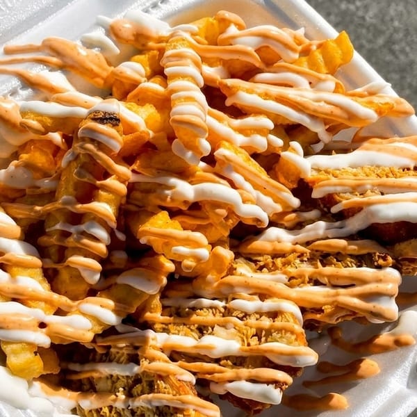 Crispy french fries and chicken pieces drizzled with white and orange sauces in a white takeout container.